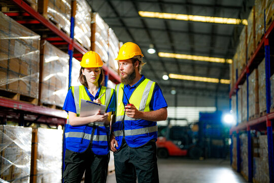 Bearded male warehouse worker and young female colleague in hard hats and safety vests shine a work light on racks during the night shift between high storage racks stacked with brown carto.