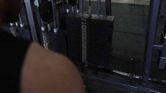Muscular man adjusting the weight pin on a gym machine stack before his workout. Male athlete preparing for strength training exercise.