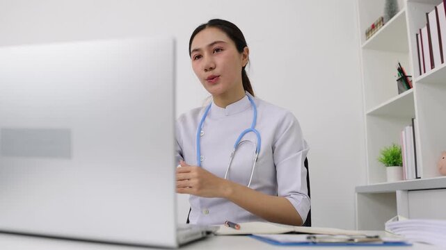 Asian female doctor providing online video consultation. Young Asian female doctor with a stethoscope providing medical advice to a patient through a video call on her laptop in a clinic office.