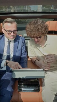 Vertical video of middle aged Caucasian businessman in suit showing digital tablet to young colleague in back seat of luxury car. Ideal for corporate travel and mobile office concepts