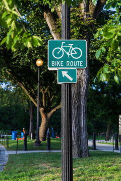 View of a green bike route sign with a directional arrow on a pole in Valley Forge National Historical Park, with trees and a park path in Valley Forge, Pennsylvania, United States.