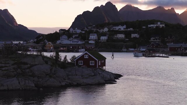 Traditional wooden rorbu cabin stands on rocky shore overlooking coastal bay and mountainous peaks in Reine, Lofoten, Norway at dawn.