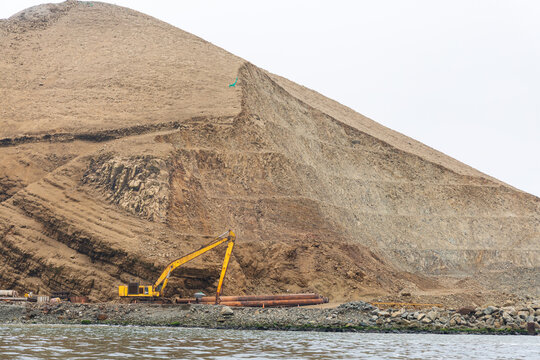 View of a yellow excavator working at the base of a large, terraced, arid hill near the coast in Chancay, Lima Region, Peru.