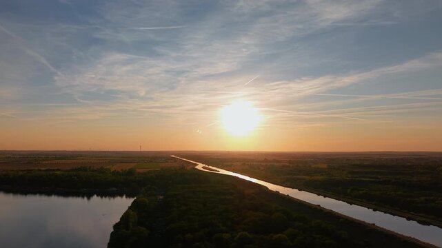 Sunset aerial panorama over Mittellandkanal in Saxony-Anhalt, Germany, with glowing water winding through flat countryside. Warm evening light reveals farmland, forest, horizon and distant turbines