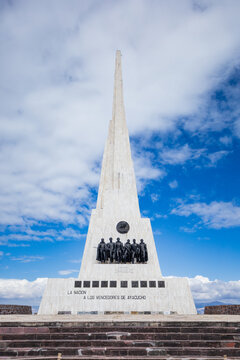 View of the Pampa De La Quinua monument, a tall white obelisk commemorating the Battle of Ayacucho, set against a bright blue sky with clouds in Quinua, Ayacucho, Peru.