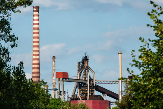 View of the industrial blast furnace and tall chimneys of the steelworks plant framed by green trees under a clear blue sky in Dabrowa Gornicza, Silesian Voivodeship, Poland.