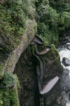 View of the stone stairs winding along the rocky cliffside at Pailon del Diablo waterfall, surrounded by lush vegetation and mist near the river in Banos, Tungurahua, Ecuador.