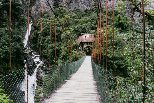 View of a wooden suspension bridge leading to a house nestled in lush forest with a waterfall in Banos, Tungurahua, Ecuador.