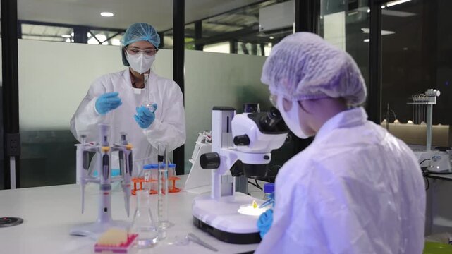 Scientists working together in a research laboratory. Two female researchers in protective suits working in a laboratory. One scientist is using a microscope while her colleague is mixing chemicals.