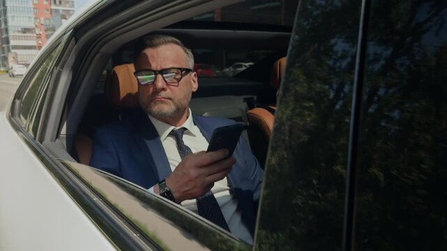 Portrait of middle aged Caucasian man in suit sitting in back seat of luxury car, holding smartphone and looking at camera. Clip is ideal for corporate travel and executive lifestyle commercials