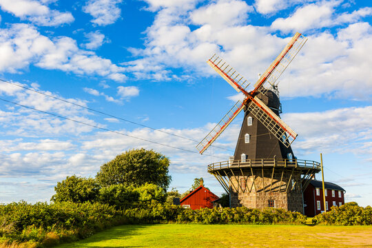 View of the historic Sardal windmill with its stone base and red blades set in a lush green landscape under a bright blue sky in Sardal, Halland County, Sweden.