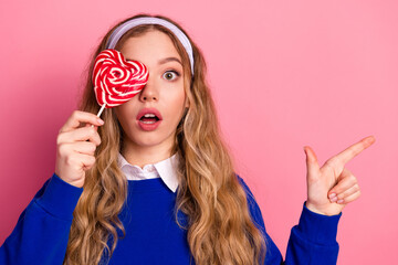Fototapeta premium Young woman with heart lollipop covers one eye and points to the side against pink background wearing blue sweater and white collar headband