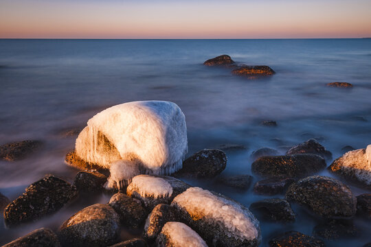 View of ice-covered rocks at Nasbokrok nature reserve during golden hour with calm sea and orange sky in Asa, Hallands lan, Sweden.