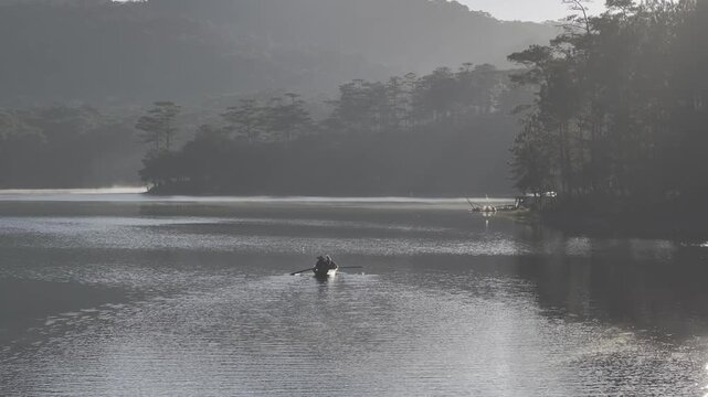Aerial drone view of a fisherman rowing across Tuyen Lam Lake at sunrise near Suoi Tia in Xuan Huong, Da Lat, Vietnam, surrounded by peaceful pine forests.