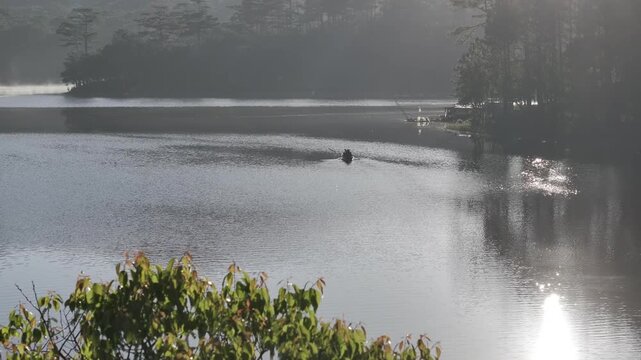 Aerial drone view of a fisherman rowing across Tuyen Lam Lake at sunrise near Suoi Tia in Xuan Huong, Da Lat, Vietnam, surrounded by peaceful pine forests.