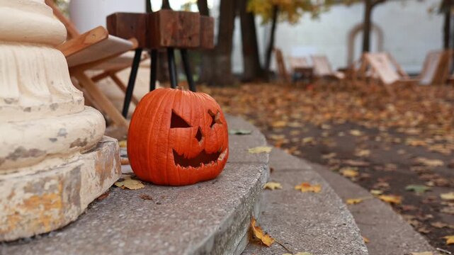 Halloween pumpkin lantern with carved face sitting near a vintage column outdoors. Seasonal fall decor concept, autumnal atmosphere, courtyard background with yellow foliage and lounge chairs.