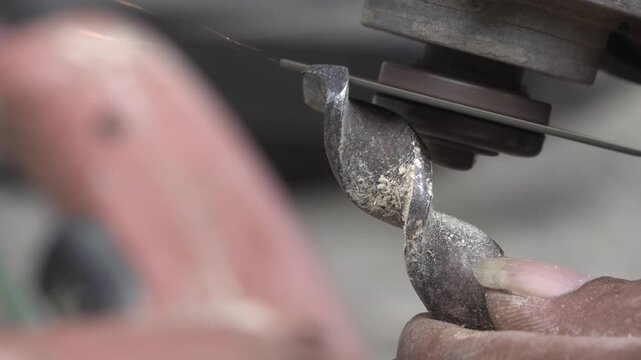 Detailed close-up view of hands carefully holding a metal auger drill bit against a spinning grinder disc to reshape and sharpen its cutting tip.

