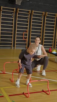 Vertical video of Caucasian kids jumping over hurdles with Caucasian female coach supervising in gym. Footage is perfect for promoting youth sports programs and physical education