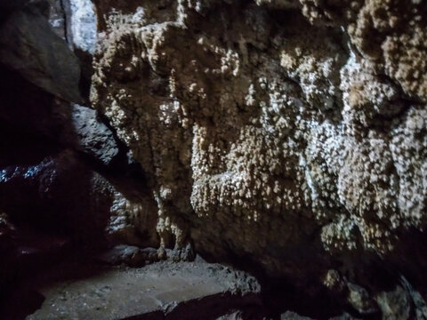 Natural limestone cavern walls display intricate white mineral formations and rugged dark rock textures captured within a mysterious underground cave illuminated by soft ambient natural light
