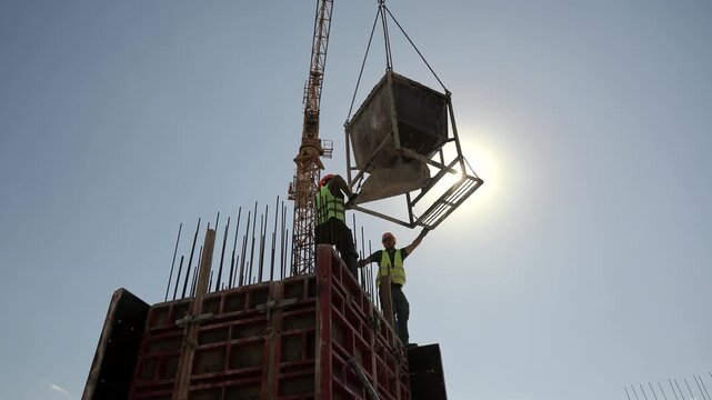 Construction workers guide suspended concrete hopper above reinforced formwork at building site, Tower crane lifts heavy equipment during active concrete work in bright sunlight.