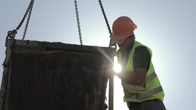 Construction worker handling suspended concrete hopper under strong backlight at building site, Safety gear and lifting chains highlight active industrial work in bright sunlight.