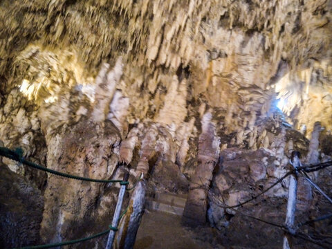 Massive stalactites and stalagmites create dramatic natural formations inside this dimly lit underground cavern featuring rugged stone textures and visible rope barriers for tourist safety