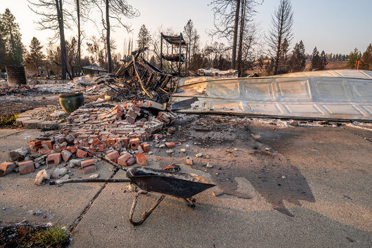 View of charred residential remains with scattered bricks, a burnt wheelbarrow, and scorched trees in the background following a wildfire in Spokane, Washington, United States.