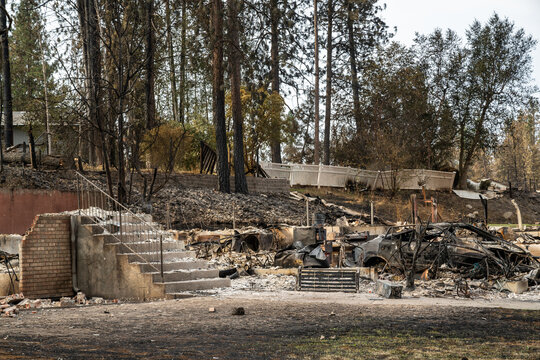 View of the charred remains of a residential property with a brick staircase and destroyed vehicle surrounded by scorched trees after the 2026 wildfires in Spokane, Washington, United States.