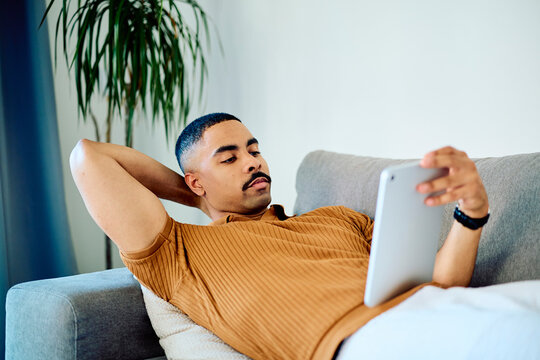 Young man with mustache lounging on a comfortable sofa at home, watching content or browsing internet on a touchscreen device