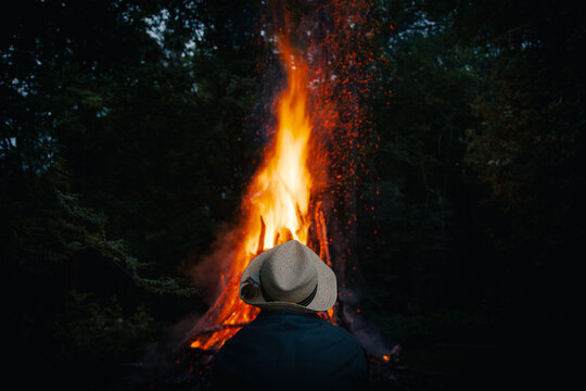 View of a person in a straw hat watching a large bonfire in a dark forest at night in Laane County, Estonia.