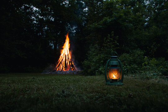 View of a large bonfire burning in a grassy field at night with a glowing green lantern in the foreground, Laane County, Estonia.