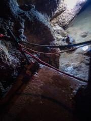 Fototapeta premium Weathered ropes tied securely to craggy limestone walls inside a dark damp cavern illuminated by soft natural light filtering from the surface above the ground