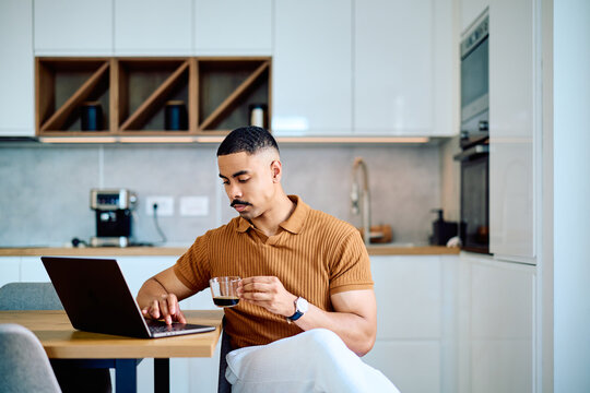 Young man telecommuting in a modern kitchen, seated at a table with laptop, sipping coffee while working and browsing online files and email