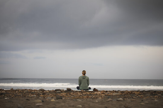 View of a person sitting in a meditative pose on the dark sand beach facing the ocean under an overcast sky in Ubud, Bali, Indonesia.