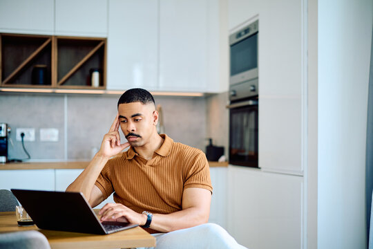 Focused man in modern kitchen working on laptop. Representing remote work, concentration, thinking, and home office lifestyle