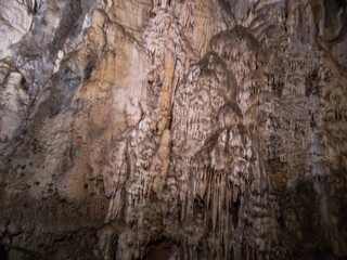 Massive limestone cave walls display intricate natural formations including stunning stalactites and detailed mineral textures captured in dim lighting within this ancient underground rocky cavern  © Graphixa