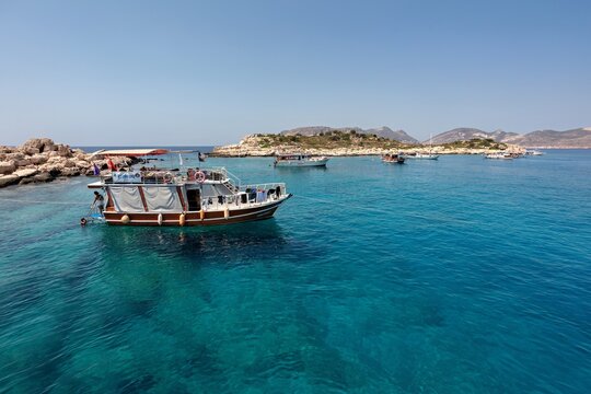 View of a wooden dive boat anchored in the clear turquoise waters of the Mediterranean Sea with rocky islands and a clear blue sky in Kas, Antalya, Turkey.