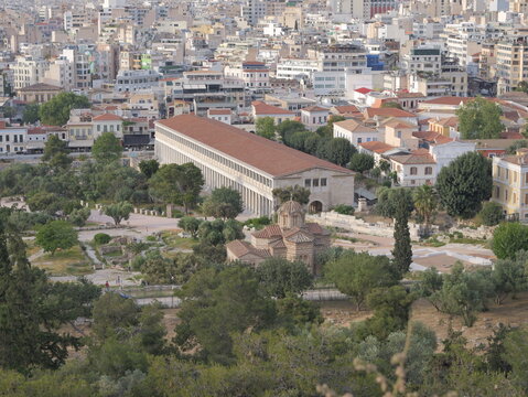 View of the Stoa of Attalos with its long colonnade and red-tiled roof, situated near a small Byzantine church amidst lush green trees and the urban cityscape of Athens, Greece.