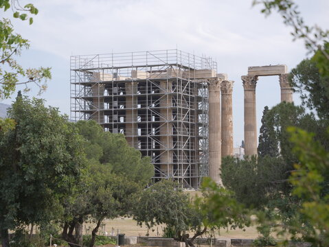 View of the Temple of Olympian Zeus surrounded by metal scaffolding during restoration work, framed by lush green trees in Athens, Greece.
