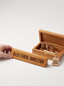 Hand holding wooden director nameplate next to box of miniature office belongings, representing employee offboarding and professional farewell concept