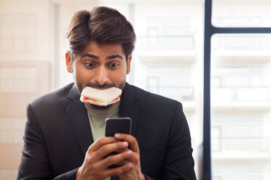A YOUNG MAN HURRIEDLY WORKING ON MOBILE WHILE EATING SANDWICH