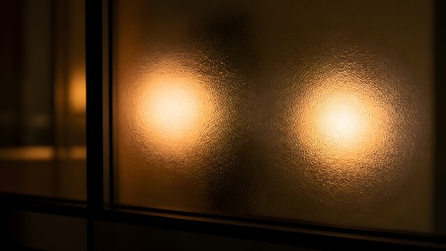 Two warm, abstract light sources seen through frosted glass pane at night, creating a soft bokeh effect and blurred reflection on a window sill.