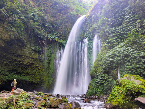 View of Tiu Kelep Waterfall cascading down a lush cliffside with a hiker standing on the rocky bank in Bayan, West Nusa Tenggara, Indonesia.