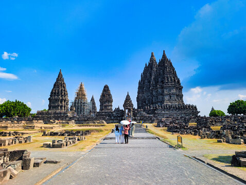 View of Prambanan Temple with tourists walking along the stone path under a bright blue sky in Kecamatan Prambanan, Daerah Istimewa Yogya, Indonesia.
