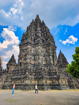 View of Prambanan Temple with intricate stone carvings and towering spires under a blue sky, with two women walking in Kecamatan Prambanan, Daerah Istimewa Yogya, Indonesia.