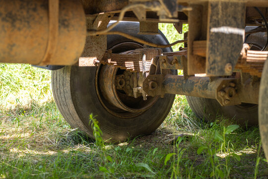 Undercarriage view of heavy vehicle trailer showing leaf spring suspension, rusty axle, and rubber tires on grassy ground in bright daylight.