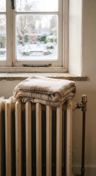 White radiator below a snowy window with folded blanket on top, pale wall, soft daylight, and simple winter heating detail
