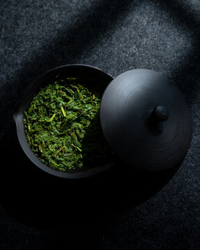 View of a black ceramic bowl filled with loose green tea leaves and its matching lid resting on a dark textured surface in the Czech Republic.