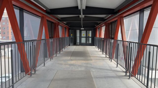 Empty pedestrian overpass bridge interior with metal structural beams, perspective view looking towards doors.