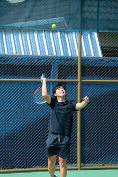 Young Asian Man Serving Tennis Ball on Outdoor Court During Sunny Day